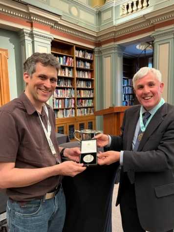 Two white men looking into the camera, between them thea silver medal in a box. Both men hold their hands on the box. In the background shelves with books (old library).